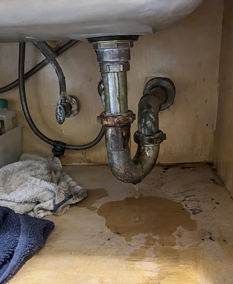 a corroded, leaking metal P-trap under a sink with a puddle on the cabinet floor