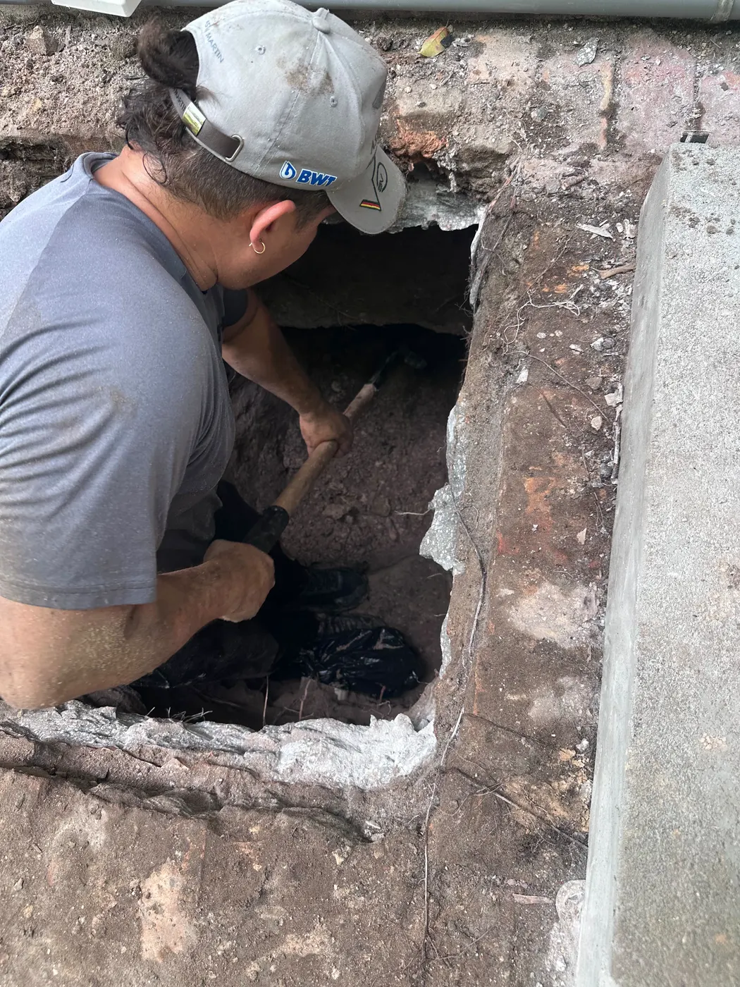 The God Plumber technician kneeling in a hand-dug excavation beside a brick wall, working on the sewer connection
