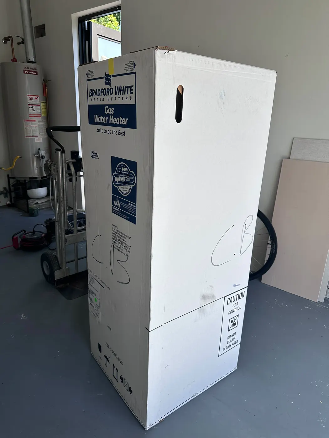 A boxed gas water heater standing in a garage next to the old unit before installation